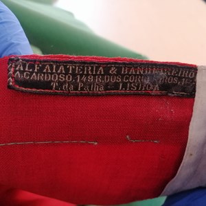 Etiqueta da bandeira (Alfaiataria e bandeireiro A. Cardoso, 149, Rua dos Correeiros, Travessa da Palha, Lisboa). António Cardoso foi o fundador desta loja que continua a produzir bandeiras, na baixa de Lisboa, com o nome «Primeira Casa das Bandeiras».